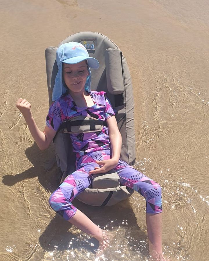 Thelita in the Bath and Beach chair, sitting in shallow sea water in a sun hat, one arm raised.