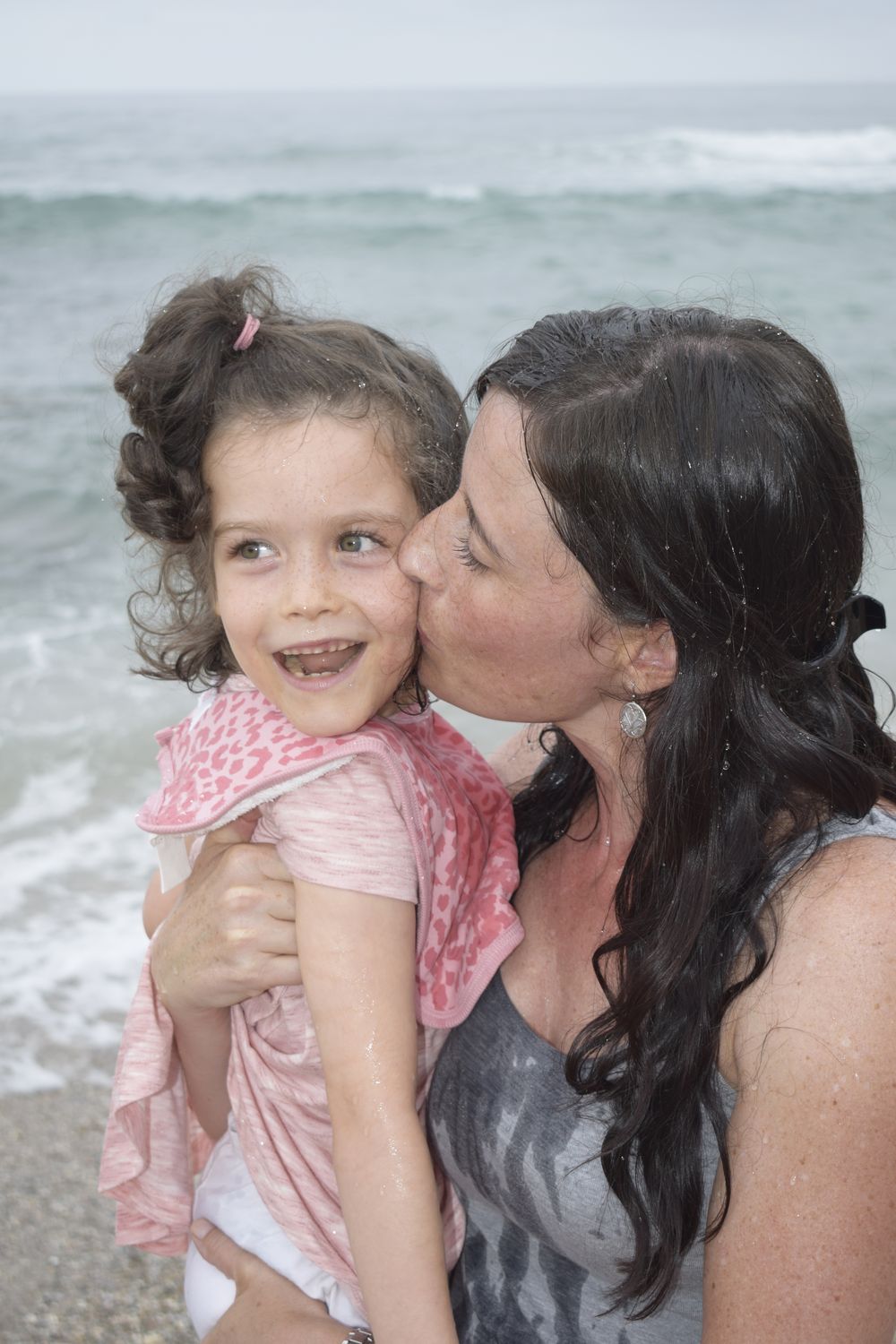 Letanea kissing Thelita on the cheek at the sea. Thelita is laughing, both of them wet from the water.
